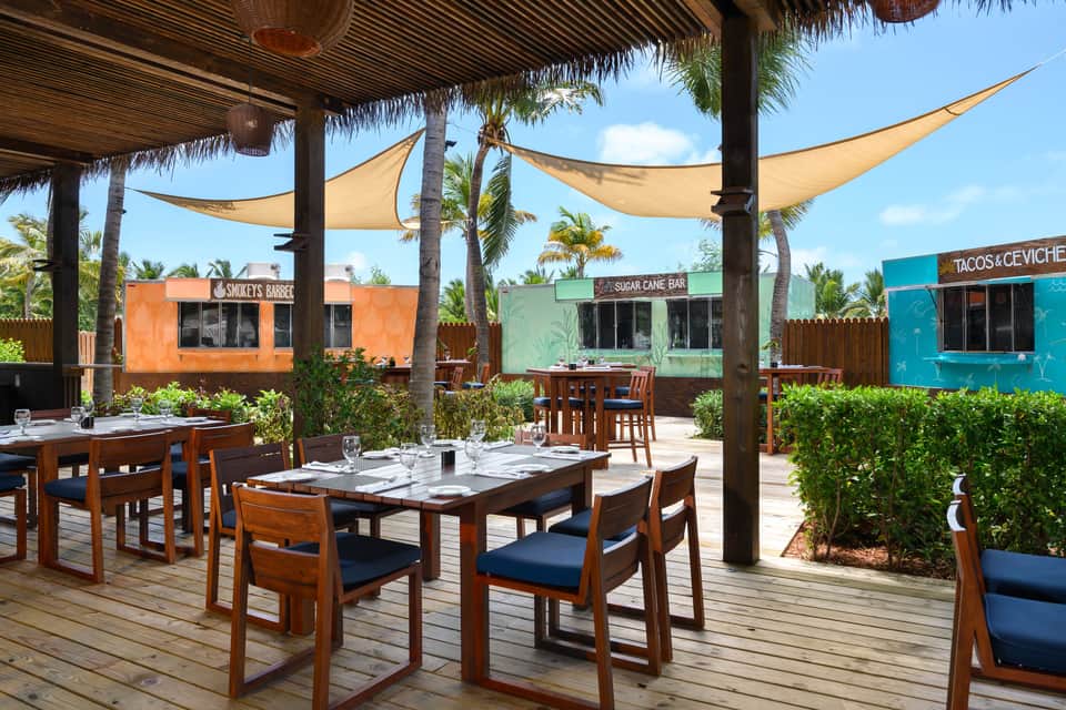 Covered patio dining with wooden tables, colorful food stalls, and palm tree shade sails