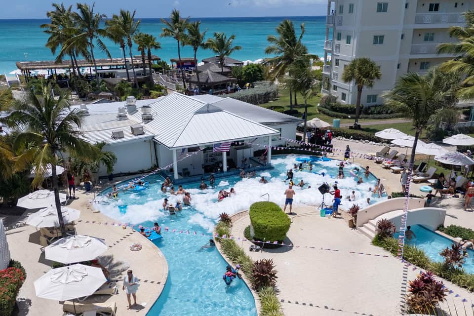 Aerial view of beachfront resort pool complex with wave pool, white umbrellas, palm trees, and turquoise ocean beyond