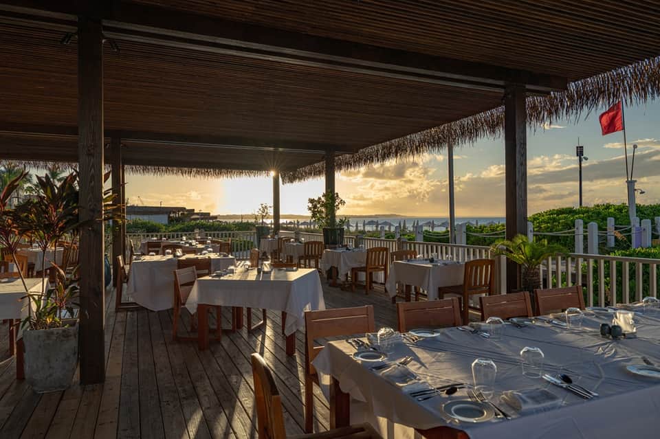 Waterfront dining terrace with white tablecloths, wooden chairs, thatched roof, and scenic sunset mountain views