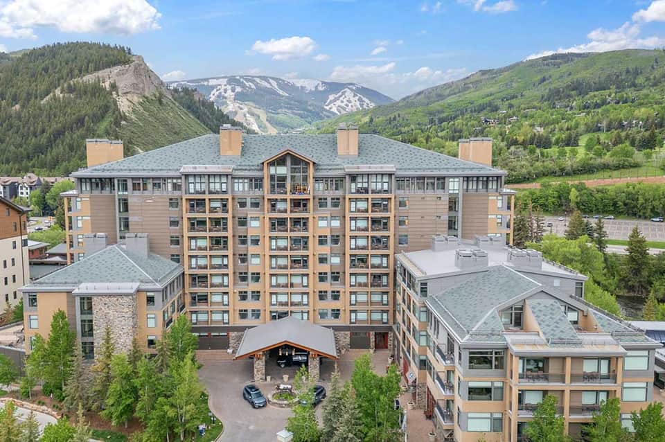 Mountain resort exterior with multi-story wood and stone building, snow-capped peaks, forested slopes, and valley backdrop