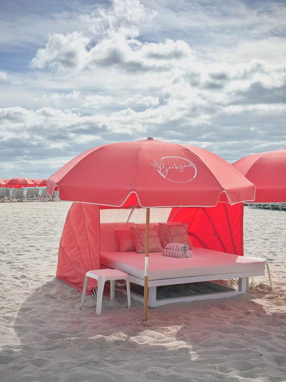 Pink cabana tent on beach with lounge bed, umbrella, pillows, and clouds visible in sky