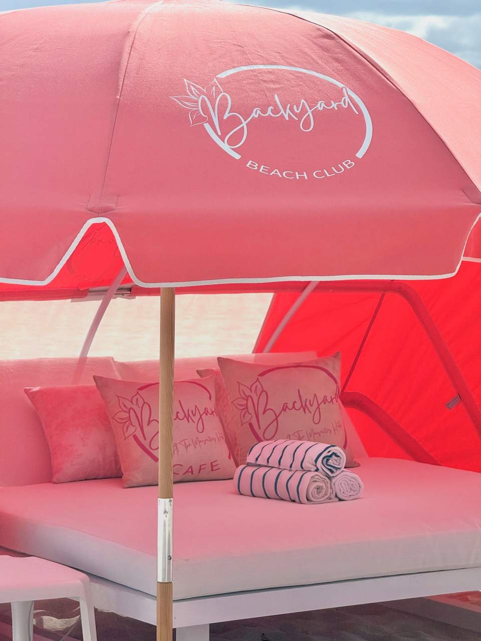 Pink cabana bed with Backyard Beach Club umbrella and lounge seating on sandy beach