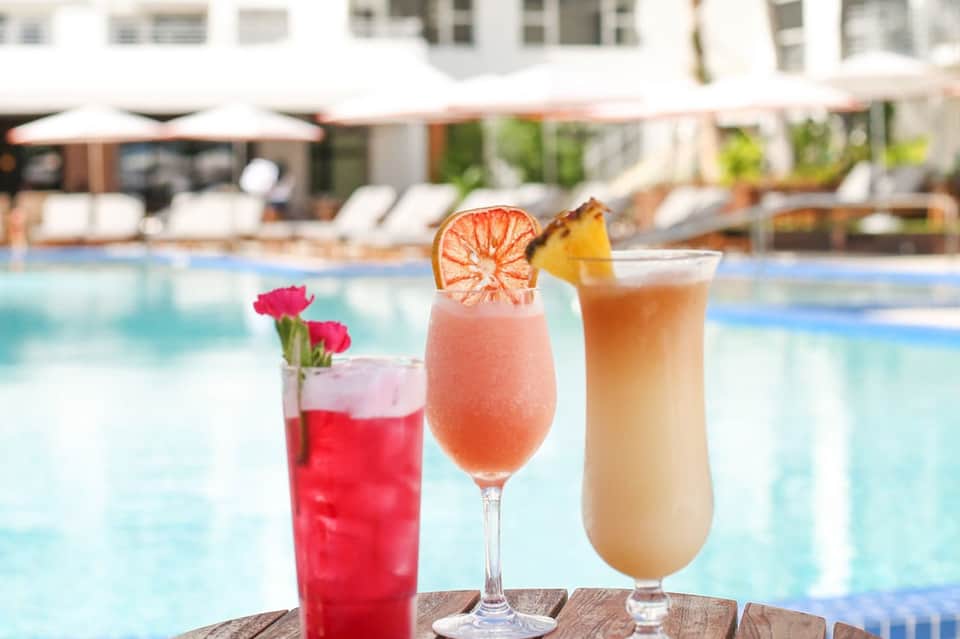 Three colorful cocktails by poolside: red drink with flower, pink grapefruit cocktail, and tropical drink with pineapple