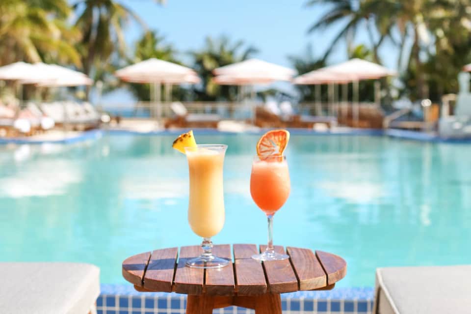 Two tropical cocktails on wooden table by resort pool with umbrellas and palm trees