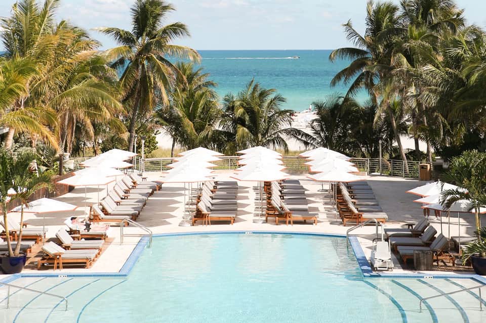 Resort pool with white umbrellas and lounge chairs facing ocean with palm trees