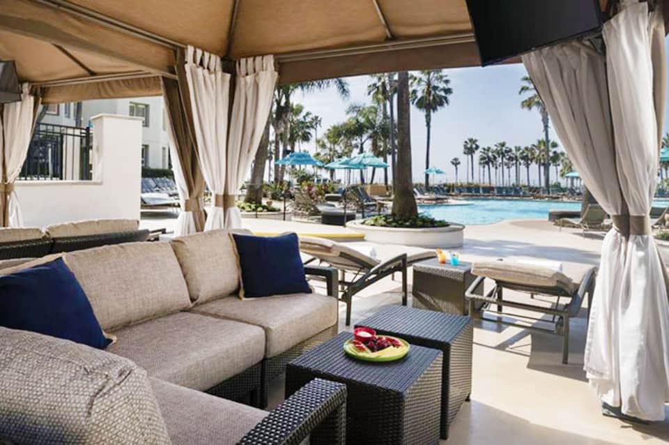 Luxury poolside lounge area with cabana, cushioned seating, ocean views, and palm trees in background