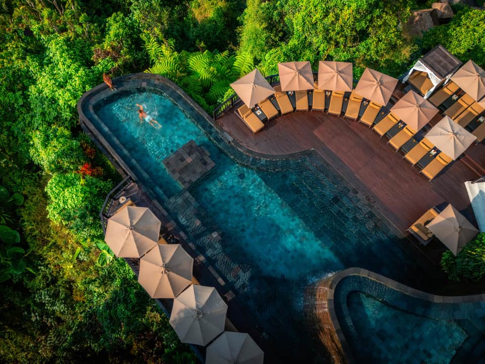 Aerial view of curved swimming pool surrounded by tan umbrellas and wooden loungers in jungle setting