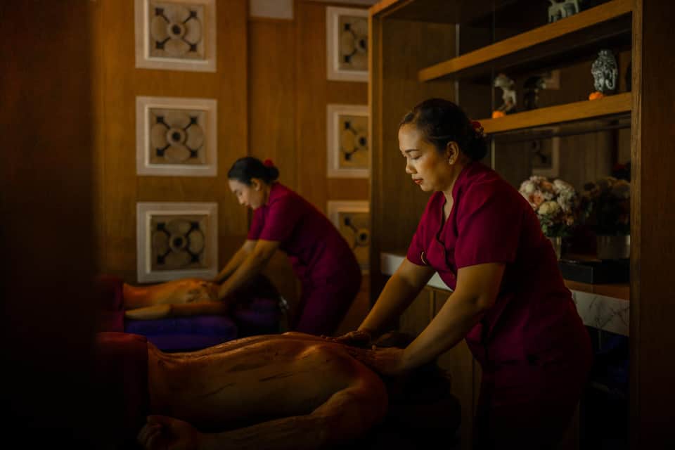 Two therapists in maroon uniforms performing massage treatments on clients in wooden spa room