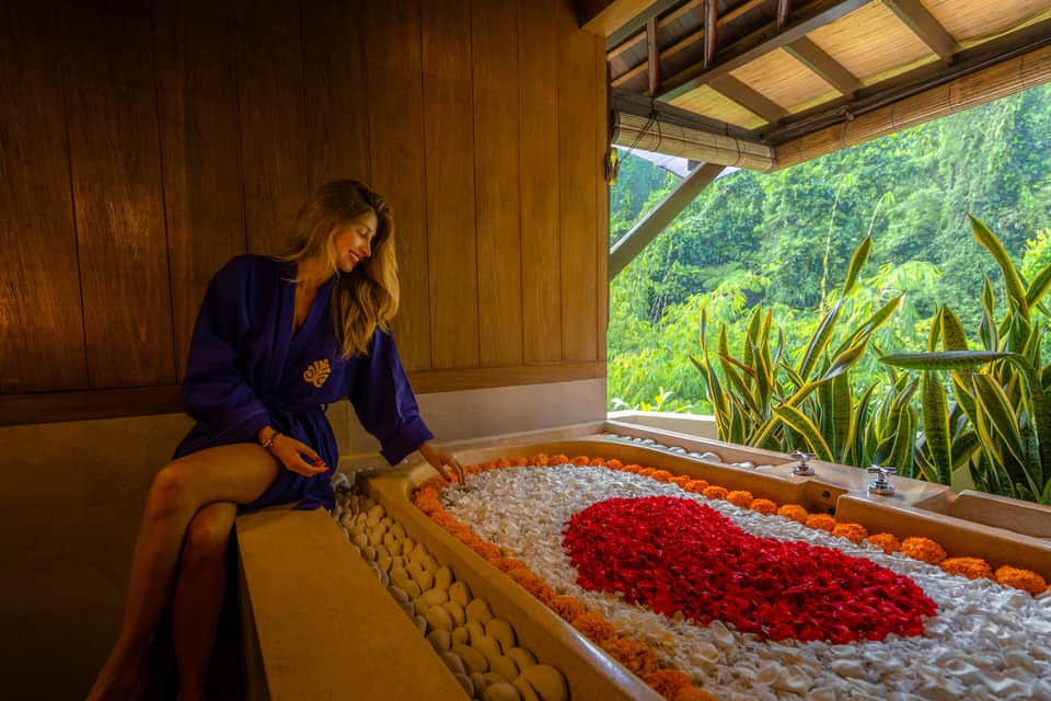 Woman in blue robe relaxing in flower petal bath with jungle mountain views through window