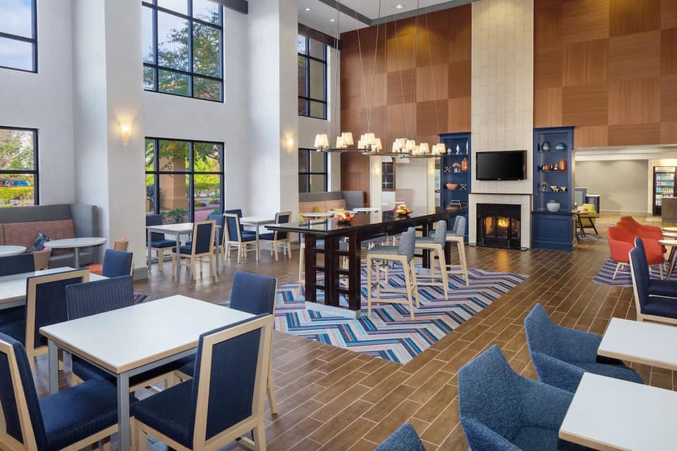 Spacious hotel lobby with high ceilings, wood accent wall, fireplace, dining area, and large windows