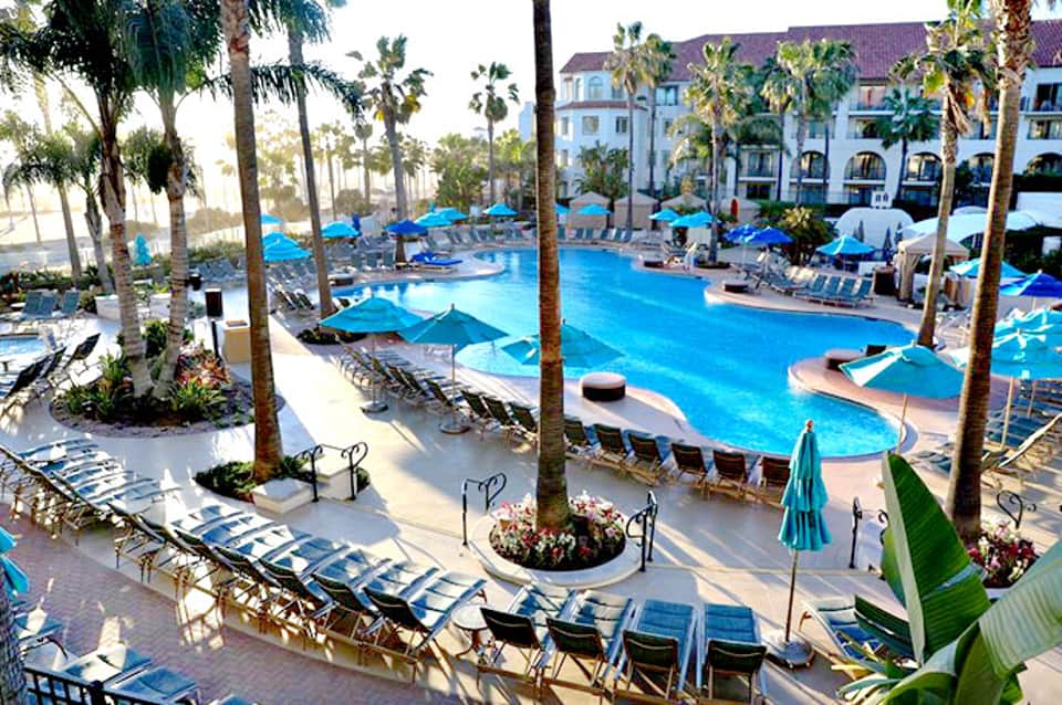 Expansive resort pool complex with blue water, palm trees, loungers, and multi-story hotel building