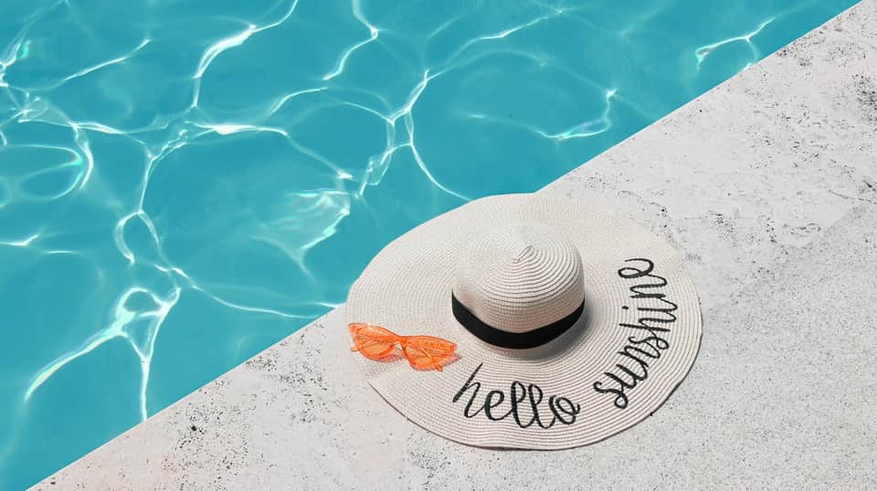 White sun hat with 'hello sunshine' text and sunglasses on pool deck next to clear water