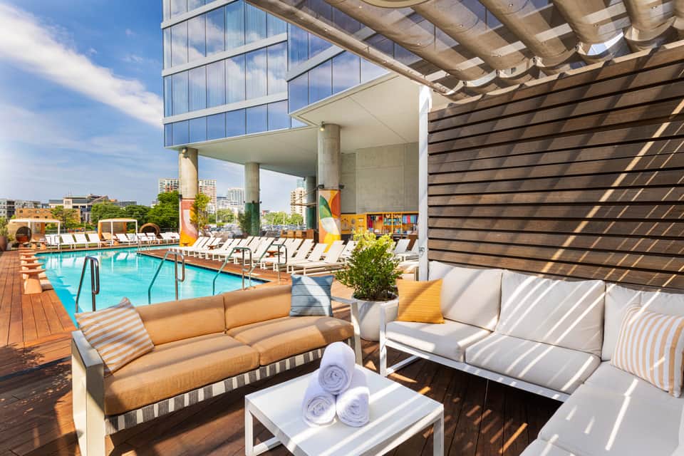 Poolside seating area with tan and white sofas, striped cushions, wooden pergola, and resort pool in background
