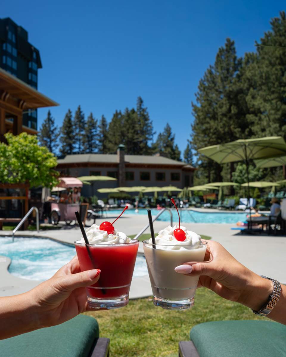 Two hands holding colorful cocktails with cherries near mountain resort pool, pine trees, and loungers