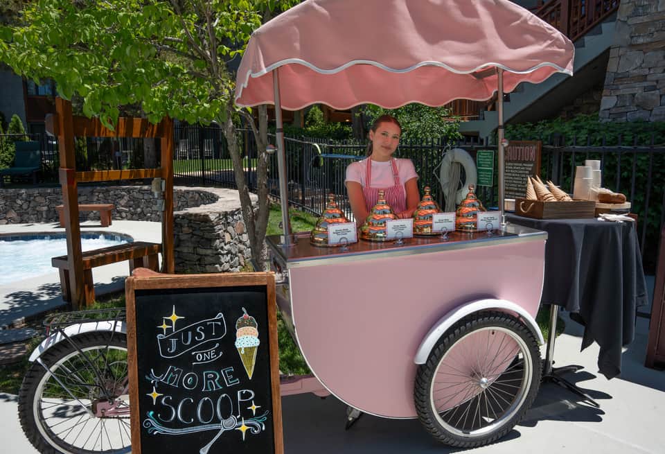 Pink ice cream cart with vendor under umbrella at resort pool area with landscaping and outdoor seating