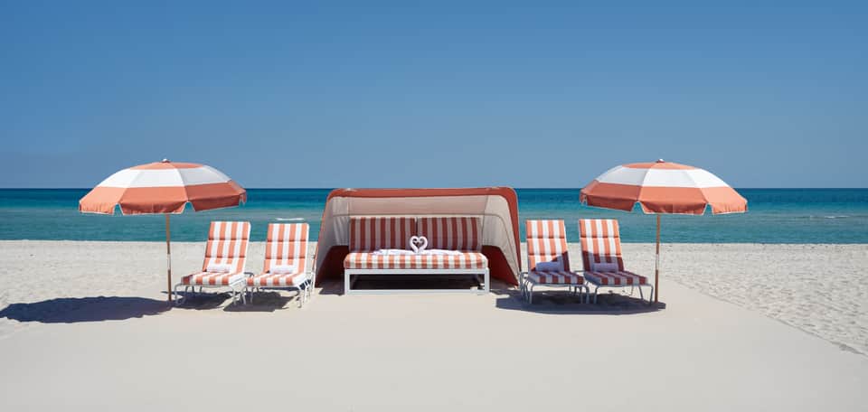 Beach setup with red and white umbrellas, striped chairs, lounge bed, and ocean view