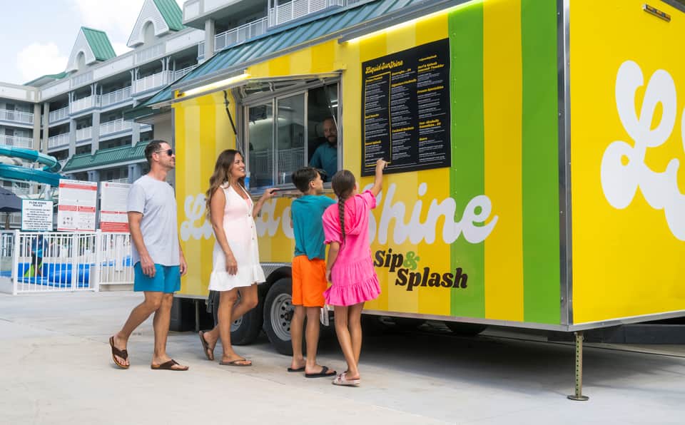 Family ordering drinks at a bright yellow food truck labeled 'Sunshine Sip & Splash' outside a resort
