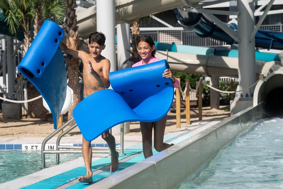 Two children carrying blue flotation devices walking toward a pool at a water park