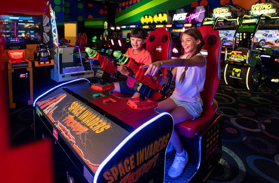 Two girls playing a red laser tag game in a colorful arcade filled with neon lighting and gaming machines
