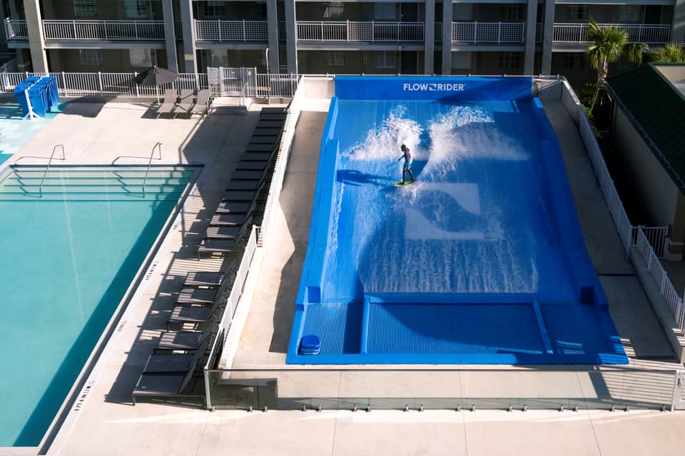 Aerial view of a surfer on the FlowRider wave machine with pool deck and resort buildings visible