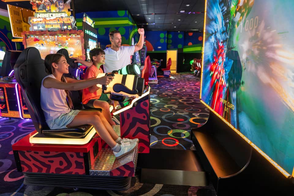 Family playing video racing game together in an arcade with neon decorations and colorful game cabinets