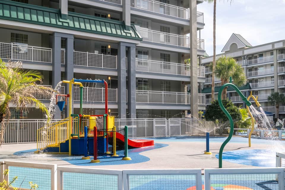 Colorful playground with yellow and green slides surrounded by resort buildings and palm trees