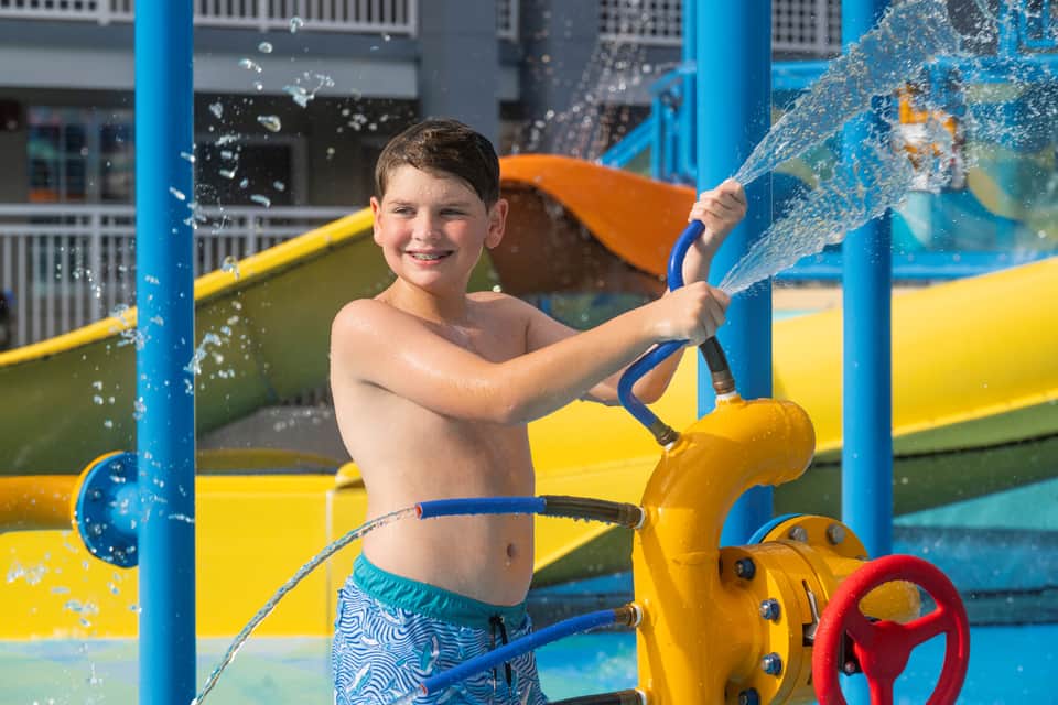Young boy at a water play structure spraying water from a blue hose, smiling at the camera
