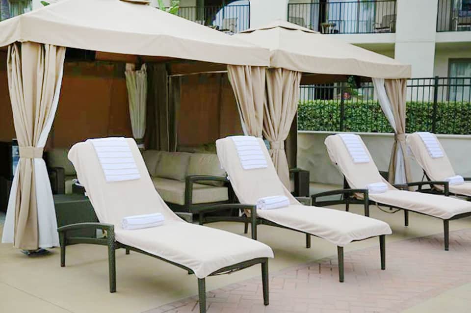 Poolside lounge area with white cushioned sun loungers under beige cabanas with draping curtains