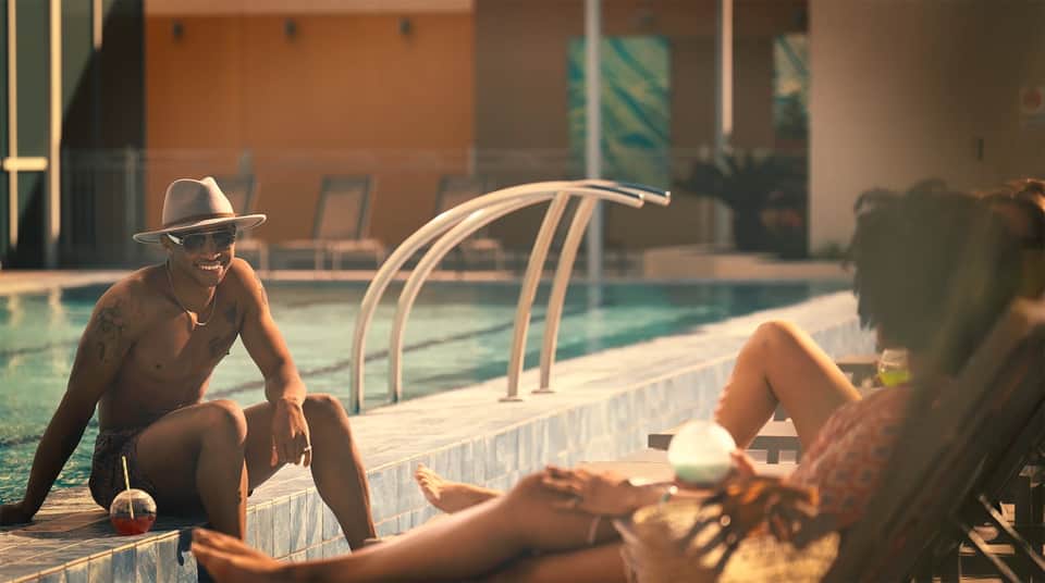 Man in hat sitting by pool edge at sunset, speaking with another guest at luxury resort pool