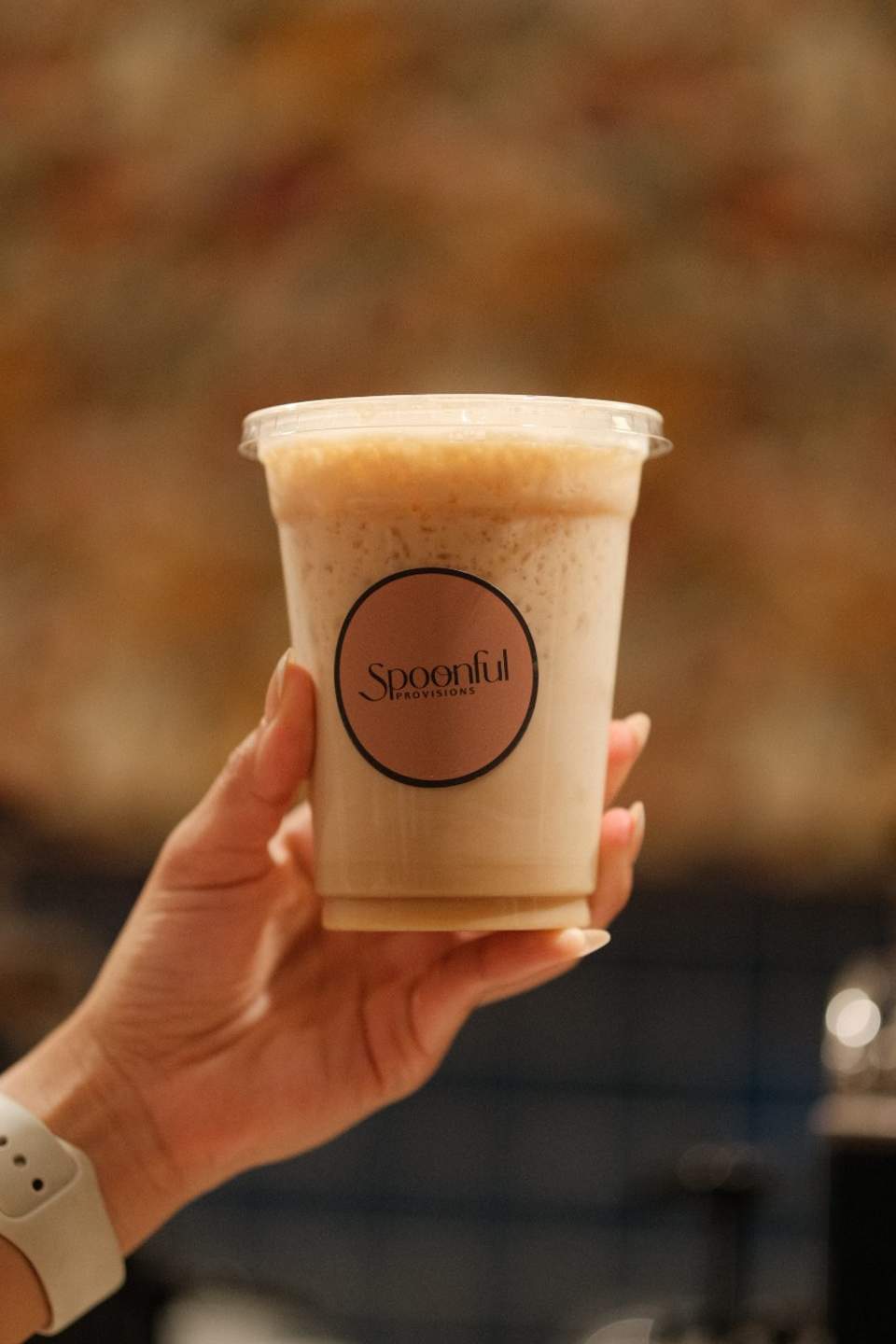 Hand holding a Spoonful Provisions branded cup of creamy beige beverage with foam top