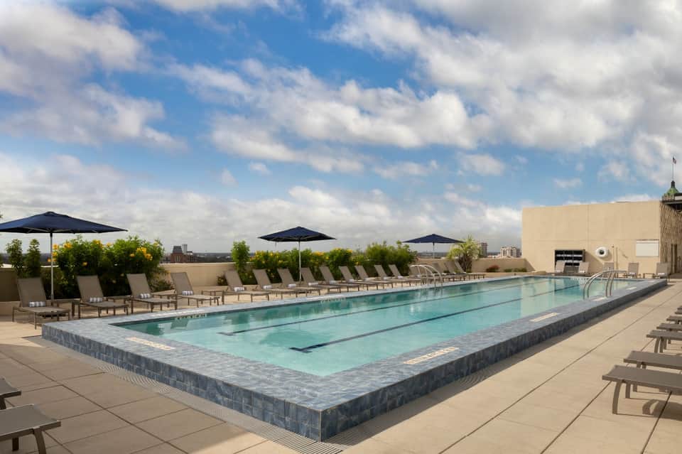 Rooftop lap pool with blue umbrellas and loungers overlooking a city skyline under cloudy skies