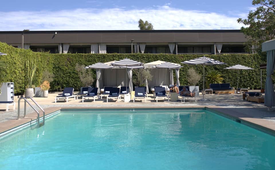 Modern hotel pool deck with striped umbrellas, white loungers, cabanas with gray curtains, and contemporary building backdrop.