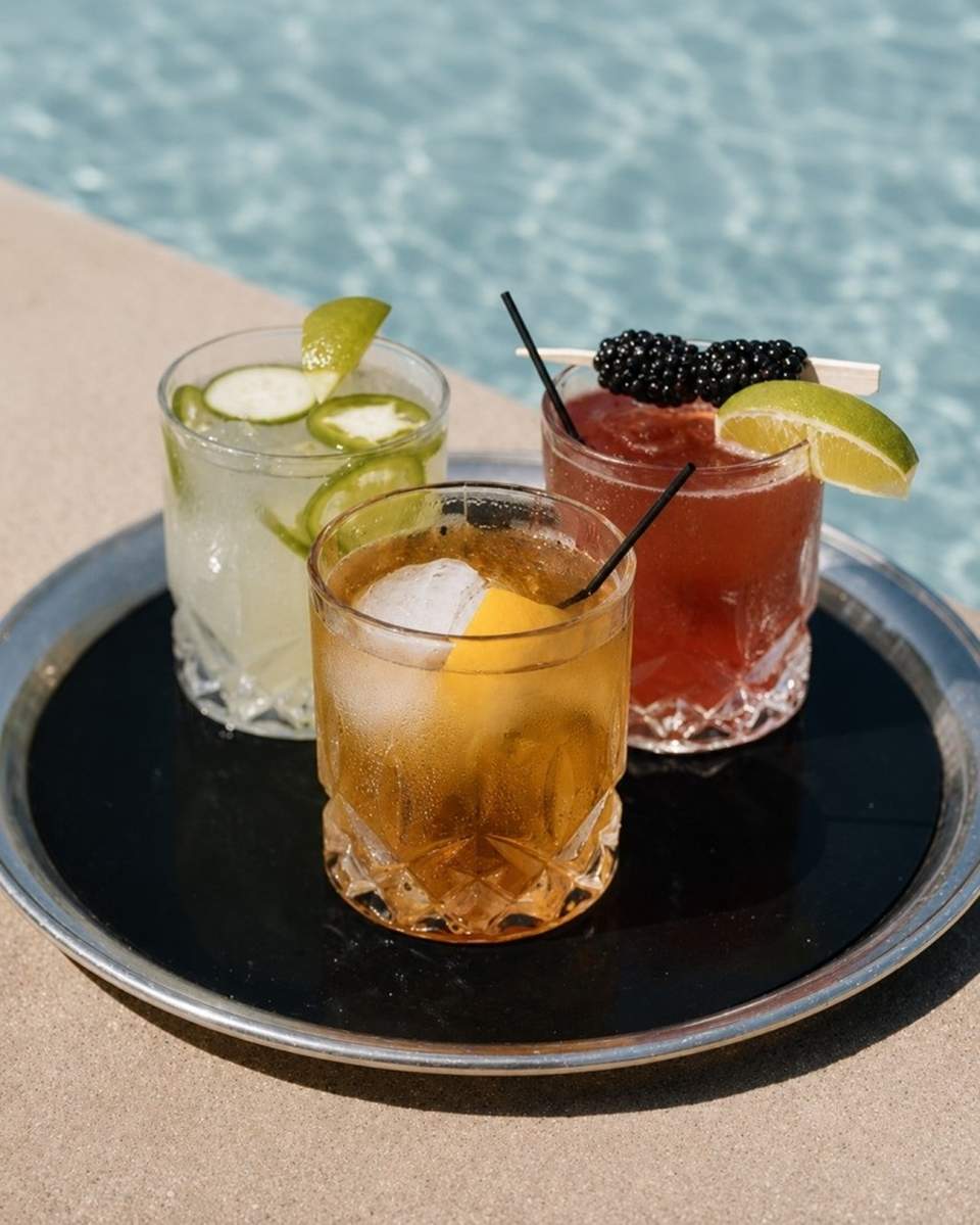 Three colorful cocktails on poolside table with lime garnish and caviar, set against sparkling blue pool water background.