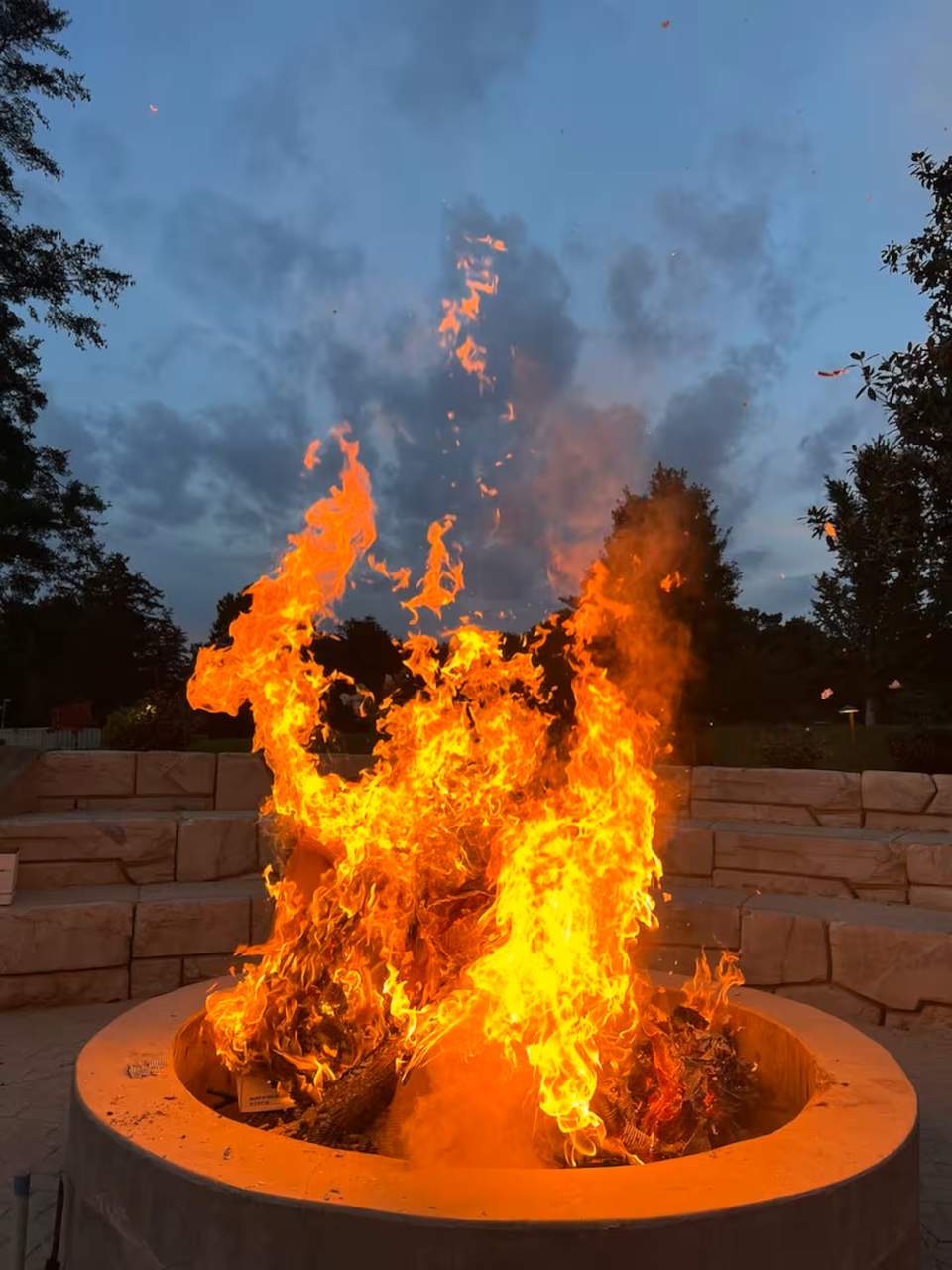 Large bonfire with bright orange flames at dusk, stone fire pit surrounded by retaining walls, trees visible in twilight.