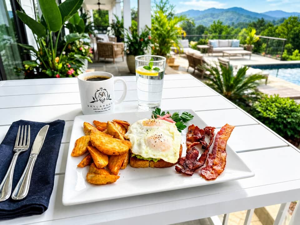 Breakfast plate with fried eggs, bacon, and potatoes on a terrace overlooking mountains and pool