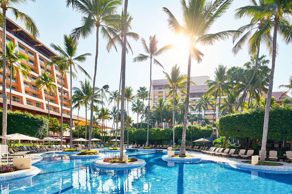 Luxurious resort pool with palm trees, lounge chairs, and orange-roofed buildings under bright sunlight