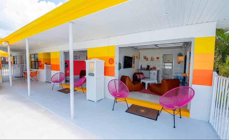 Vibrant outdoor lounge area with yellow and orange striped awning, pink wire chairs, brown bean bags, and white columns
