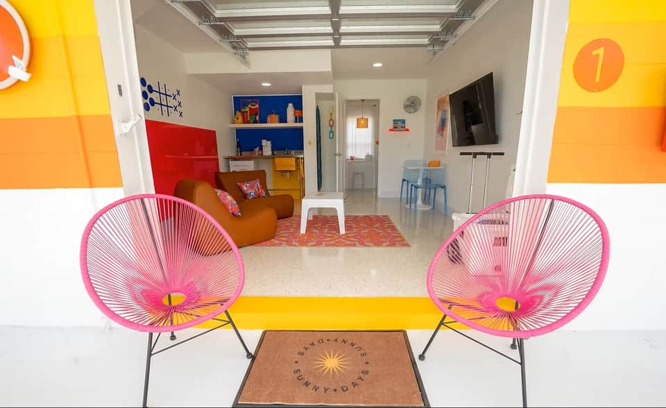 Bright modern living space with pink egg chairs on yellow stripe, orange and yellow wall panels, brown armchair, and blue shelving