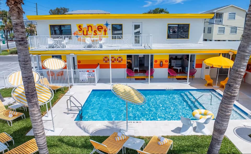Retro-style motel with bright yellow trim, pink seating visible through windows, and rectangular pool with loungers