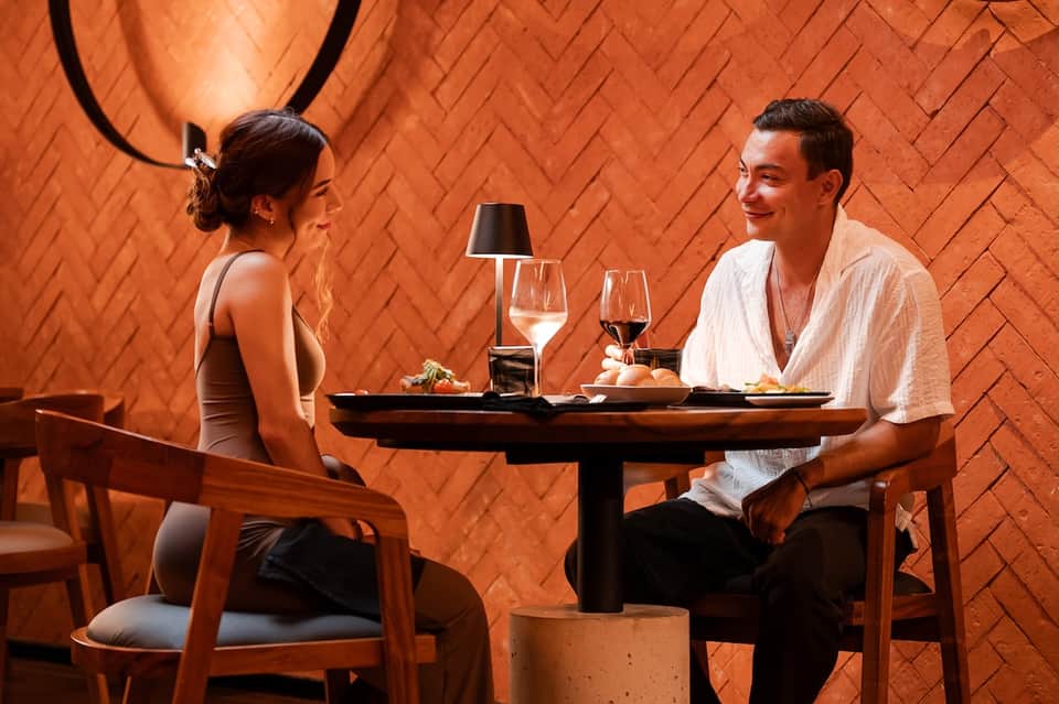 Couple dining together at an intimate table in a warm orange-brick restaurant with wine glasses and plated dishes