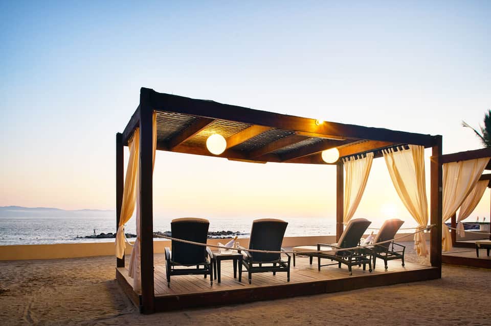 Beachfront cabana with cream curtains, lounge chairs, and ocean sunset view