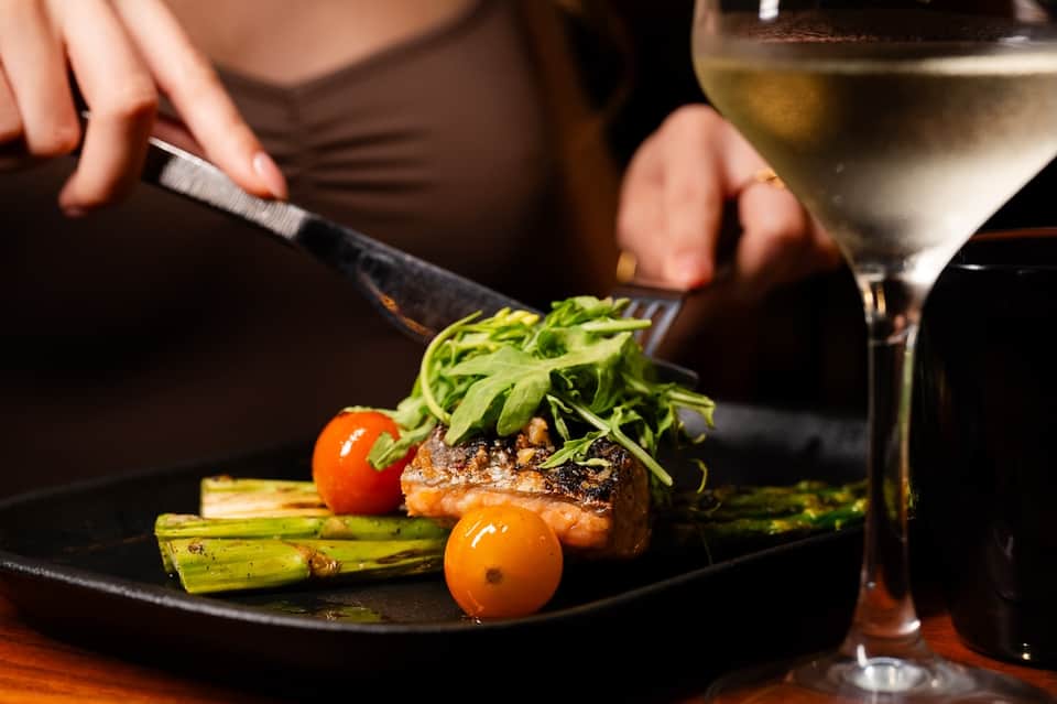 Close-up of a gourmet plated dish featuring seared meat, green asparagus, cherry tomatoes, and microgreens with wine glass