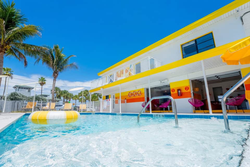 Vibrant resort pool with yellow and white floaties, pink metal chairs on deck, and colorful striped umbrellas