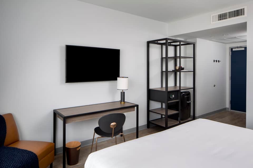 Modern hotel room with white walls, dark desk with TV, black industrial shelving unit, tan accent chair, and minimalist decor