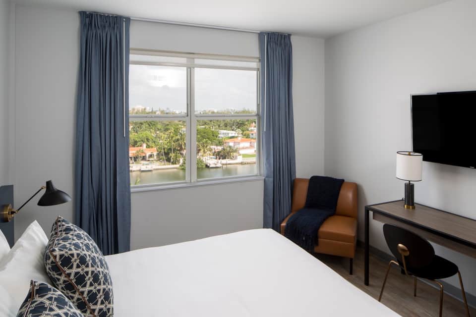 Bright hotel room with large windows overlooking water, blue curtains, white bed with patterned pillows, and tan accent chair