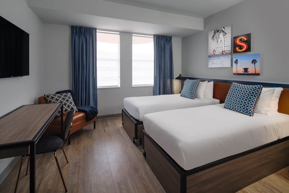Modern hotel bedroom with two queen beds, blue curtains, wooden desk, and wall-mounted TV with contemporary artwork