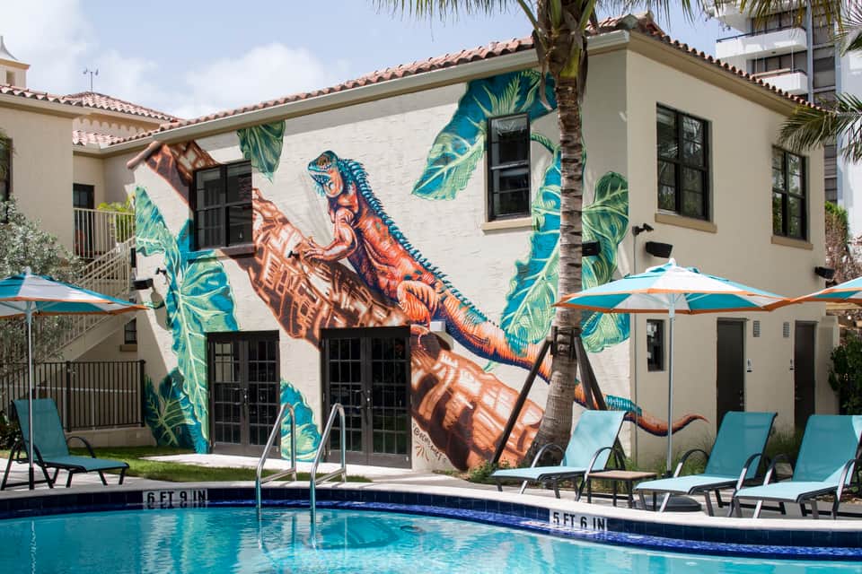 Resort pool area with colorful dinosaur mural on white building, blue lounge chairs, umbrellas, and palm trees