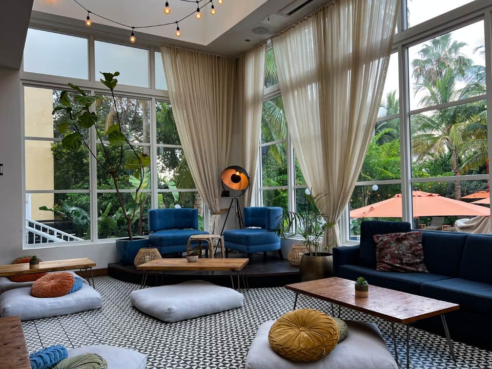 Contemporary lounge with blue curved seating, wooden tables, floor cushions, potted plants, pendant lighting, and floor-to-ceiling windows