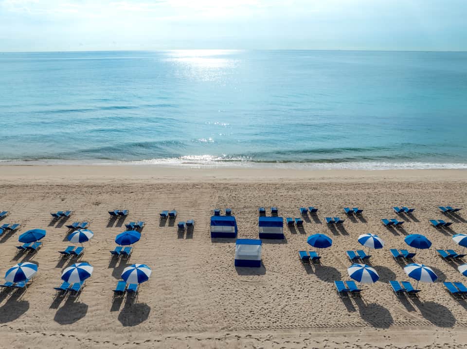 Aerial view of organized beach with blue and white umbrellas, lounge chairs, and calm turquoise ocean