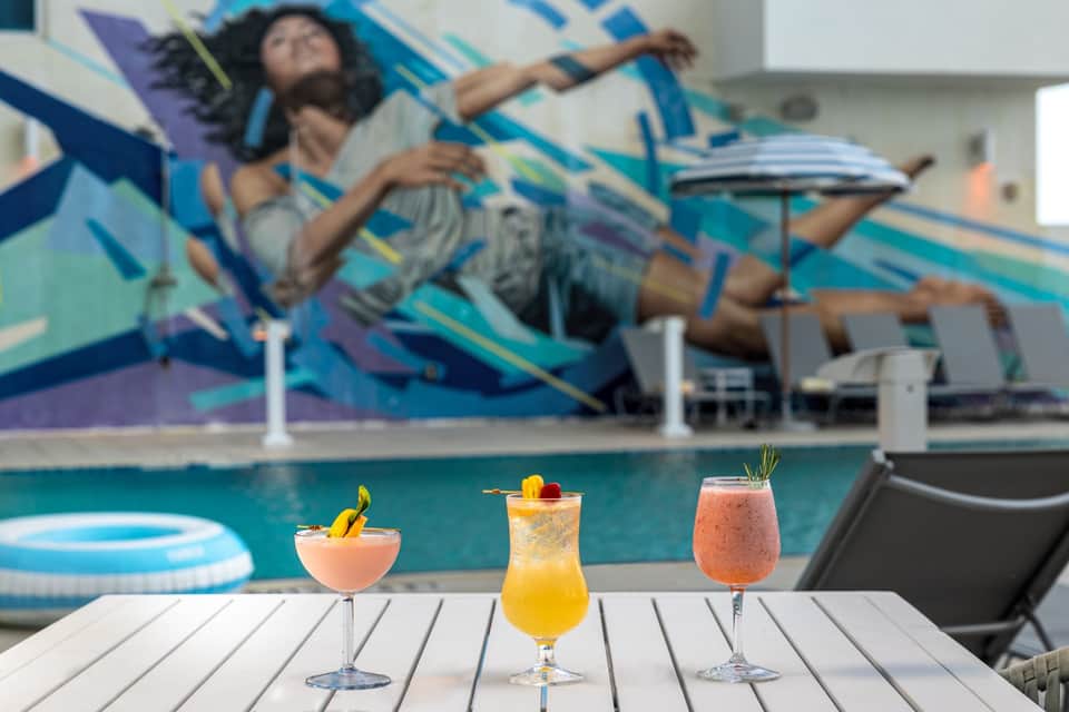 Tropical cocktails on poolside table with mural backdrop and lounging guest